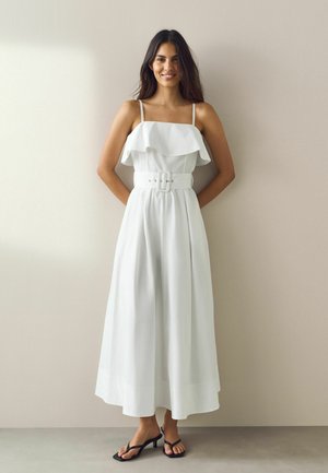 Kvinde med langt mørkt hår iført en hvid stropløs midi-kjole med bælte og sorte hælede sandaler, stående op ad en beige væg.