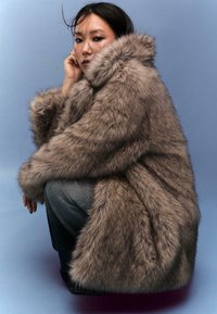 Cappotto in pelliccia sintetica marrone con colletto ampio e tessuto spesso e imponente. Il modello è accovacciato, indossando jeans grigi. Lo sfondo è di un azzurro chiaro.