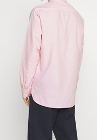 Camicia rosa chiaro a maniche lunghe con colletto, polsini con bottoni e orlo curvo. Realizzata in un tessuto liscio con una texture sottile.