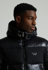 Die schwarze Puffjacke verfügt über eine glänzende Oberfläche, einen hohen Kragen, einen Reißverschluss vorne und ein Logodetail auf der Brust. Texturierte Einsätze betonen das Design.