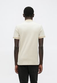 Homme aux cheveux courts et bouclés portant un polo beige à manches courtes et un pantalon noir, vu de dos sur un fond blanc uni.