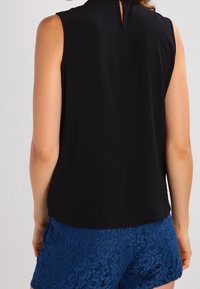 Schwarze ärmellose Bluse mit hohem Kragen, ausgestattet mit einem Schlitz hinten, kombiniert mit blauen Spitzen-Shorts, die ein florales Muster zeigen.