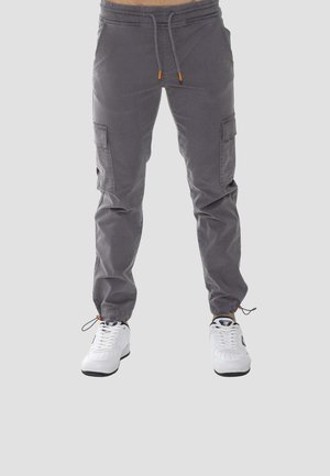 Pantalon cargo gris avec poches latérales, cordons réglables à la taille et aux chevilles, porté avec des baskets blanches sur un fond uni.