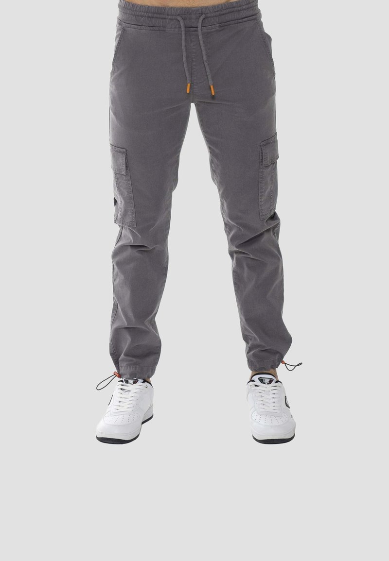 Pantalon cargo gris avec poches latérales, cordons réglables à la taille et aux chevilles, porté avec des baskets blanches sur un fond uni.