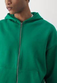 Veste à capuche verte zippée en tissu doux, dotée d'une capuche, de poches avant et d'un zip argenté. Texture lisse et coupe décontractée.