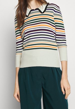 Kvinde iført en ribstrikket sweater med flerfarvede, vandrette striber og en spids krave, kombineret med mørkegrønne bukser med vide ben.