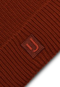 Mézelt piros kötött sapka vízszintes bordázással. Középen egy négyzet alakú barna folt található, amelyen az "J" betű van belefűzve az illeszkedő szálon.