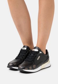Scarpe sportive nere con finitura lucida, dotate di un design con lacci, dettagli metallici e una punta decorata. Suola testurizzata visibile.