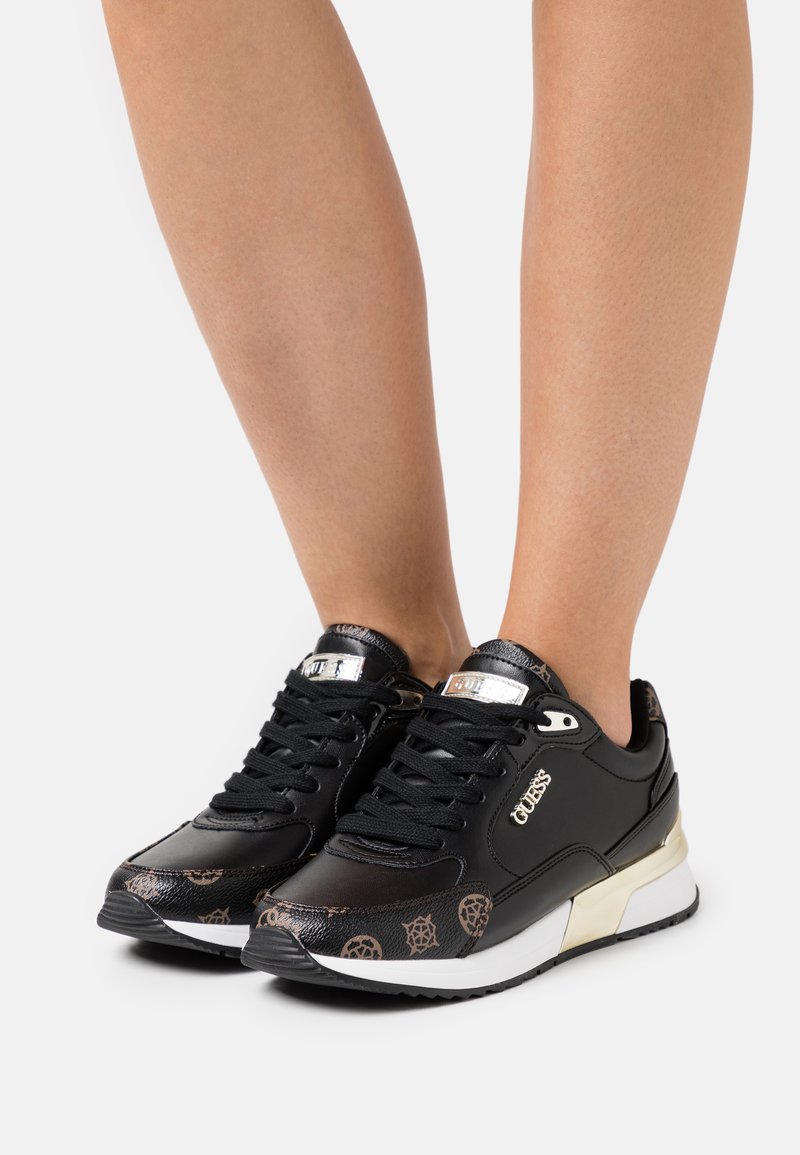 Scarpe sportive nere con finitura lucida, dotate di un design con lacci, dettagli metallici e una punta decorata. Suola testurizzata visibile.