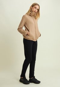 Femme aux cheveux blonds portant une veste matelassée beige, un pantalon noir et des baskets noires, se tenant les mains dans les poches devant un fond neutre.