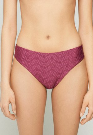 Frau trägt hochgeschnittene, strukturierte bikinihosen in mauve mit chevronmuster, gezeigt von der taille bis zum oberen oberschenkel.