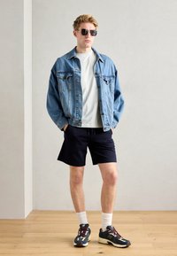 Jeansjakke, hvit t-skjorte, marineblå shorts og joggesko med fargerike detaljer. Svarte solbriller fullfører det avslappede antrekket.