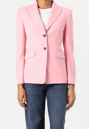 Kvinde iført en tætsiddende pink blazer med to sorte knapper og klaplommer, kombineret med blå jeans og en hvid top indenunder.