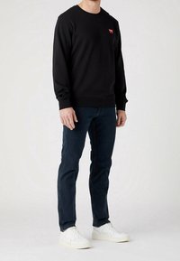 Svart bomullströja med rundad halsringning, prydd med en liten röd logotyp på vänster bröst; kombinerad med mörkblå jeans och vita sneakers.