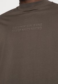 Brun t-shirt med rund hals och subtil textur; har präglad branding "CALVIN KLEIN JEANS" över bröstet. Mjuk tyg.