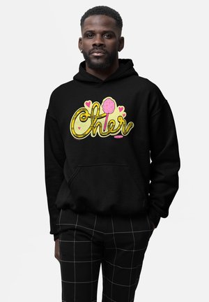 Mężczyzna w czarnej bluzie z kapturem z żółtym i różowym napisem „Cher” oraz różowymi serduszkami, w połączeniu z czarnymi spodniami z białymi liniami kratki.