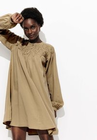 Robe longue khaki à manches longues avec un col smocké, une coupe décontractée et un ourlet fluide. Tissu doux avec un léger brillant.