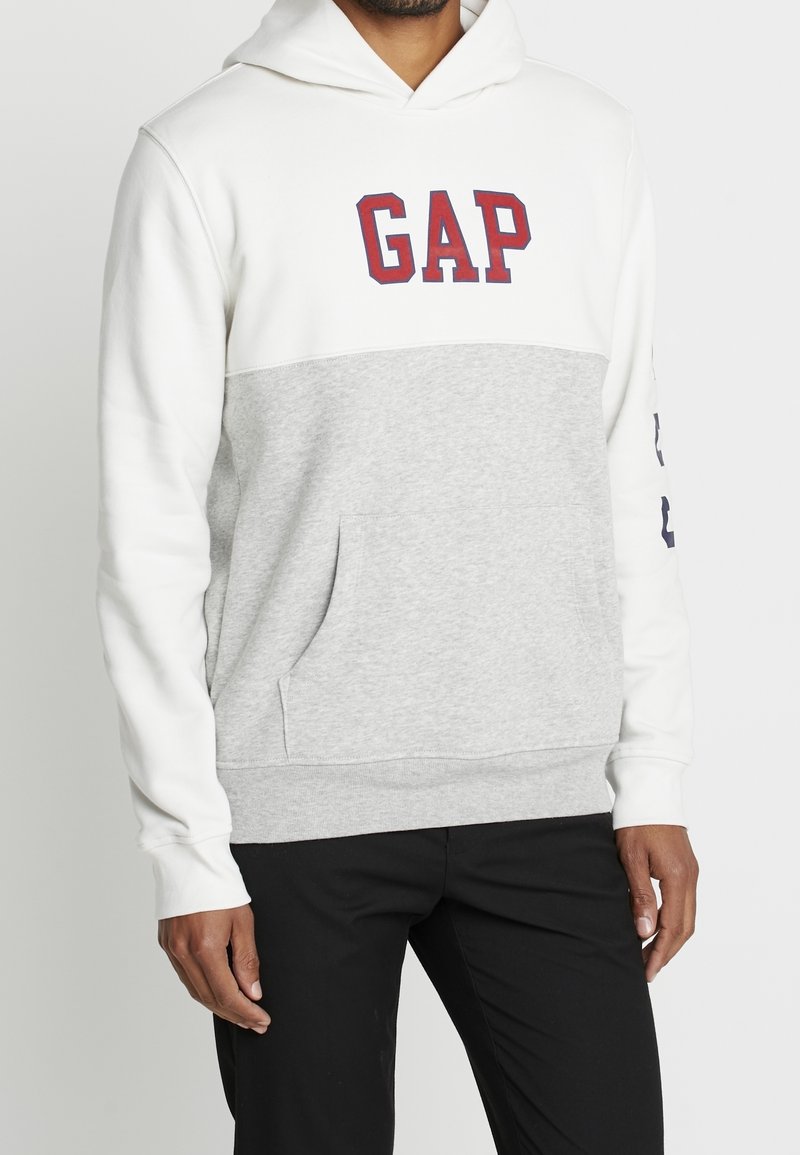 Sudadera con capucha que presenta un diseño en bloques de color blanco y gris, un bolsillo frontal y un logo "GAP" en rojo en el pecho. Material de algodón suave.