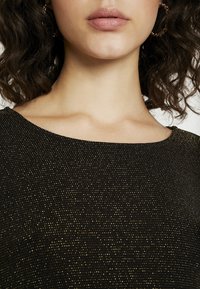 Schwarzes Oberteil mit glatter, metallischer Textur, das mit kleinen goldenen Punkten verziert ist, breitem Ausschnitt und kurzen Ärmeln; Nahaufnahme des oberen Brust- und Halsbereichs.