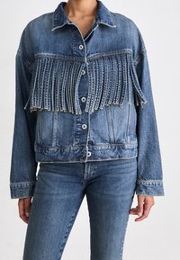 Veste en jean avec détail de franges verticales sur la poitrine, portée avec un jean bleu assorti.