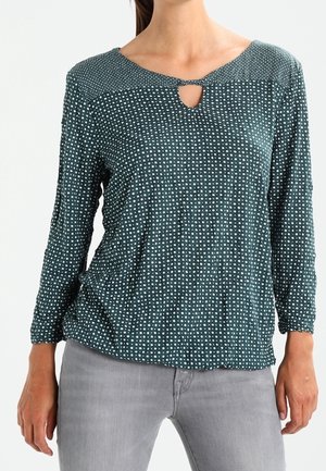 Woman wearing a dark green long-sleeve blouse with white polka dots and a small keyhole neckline, paired with light gray pants.