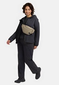 Beige Bauchtasche über einer schwarzen Jacke und Hose getragen. Die Tasche verfügt über einen Reißverschluss und einen verstellbaren Gurt, vor einem neutralen Hintergrund.