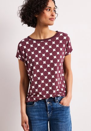 Kvinde med krøllet hår iført en vinrød og hvid mønstret kortærmet skjorte og blå jeans, stående med den ene hånd i lommen.