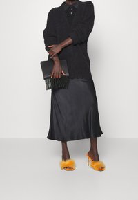 Person klädd i en svart knappad cardigan, svart satin midi-kjol, håller en svart pärlmuff, och bär orange fluffiga högklackade sandaler.