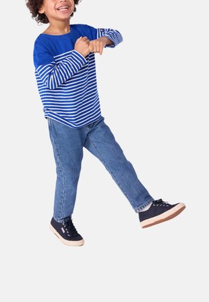 Camicia a maniche lunghe a righe blu e bianche, jeans blu e sneakers nere con suole beige. Bambino sorridente in una posa giocosa.