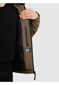 Brązowa izolowana kurtka z czarną pikowaną podszewką i kieszenią na zamek, wyposażona w metkę marki wewnątrz oraz solidne zapięcie na zamek.