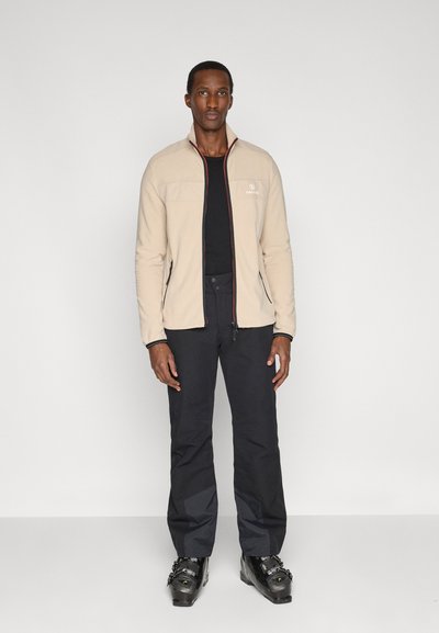 Großer Mann trägt eine beige Jacke mit Reißverschluss, schwarze Hosen und schwarze Skistiefel, steht vor einem schlichten hellen Hintergrund.