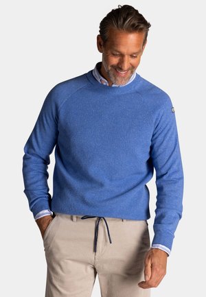 Man draagt een blauwe trui met ronde hals over een overhemd met kraag en beige broek, handen in de zakken, kijkt naar beneden en glimlacht tegen een lichte achtergrond.