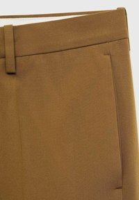 Ceinture de pantalon en tissu marron avec passant de ceinture et bord de poche latérale visible sur un fond neutre.