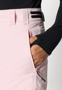 Helle rosa Skihosen mit glatter Textur, ausgestattet mit einer seitlichen Tasche mit Reißverschluss und schwarzen Klettverschlussakzenten am Bund.
