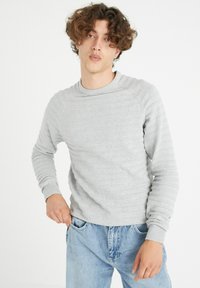 Suéter de punto gris claro con un patrón texturizado, cuello redondo y puños de canalé, combinado con jeans de mezclilla azul claro.