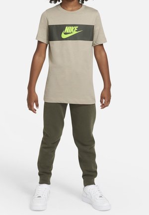 Dziecko ubrane w beżowy t-shirt Nike z zielonym logo, ciemnozielone spodnie dresowe i białe tenisówki, stojące na białym tle.