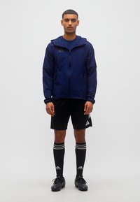 Άνδρας που στέκεται με σκούρο μπλε μπουφάν, μαύρο σορτς, μακριές μαύρες κάλτσες και μαύρα ποδοσφαιρικά παπούτσια σε απλό λευκό φόντο.