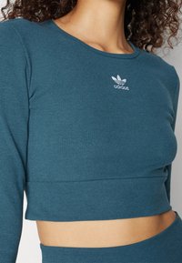 Žena v tealově zeleném žebrovaném krátkém topu s dlouhým rukávem, na němž je bílé logo Adidas umístěné uprostřed u výstřihu.