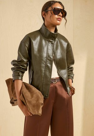 Frau trägt dunkelgrüne Lederjacke, braune Hosen mit weitem Bein, Sonnenbrille und hält eine große braune Wildleder-Clutch vor beigem Hintergrund.