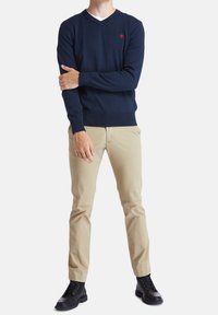 Námořnický svetr s výstřihem do V z měkkého pleteného materiálu, kombinovaný s khaki kalhotami a černými šněrovacími botami. Červený emblém na levé straně hrudi.