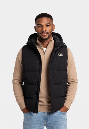 Homme portant une veste matelassée noire avec capuche par-dessus un pull zippé beige et un jean bleu clair, debout les mains dans les poches.