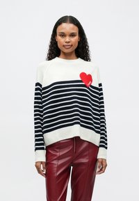 Mujer joven vistiendo un suéter a rayas blanco y azul marino con un detalle de corazón rojo y pantalones de cuero rojo, de pie contra un fondo liso.