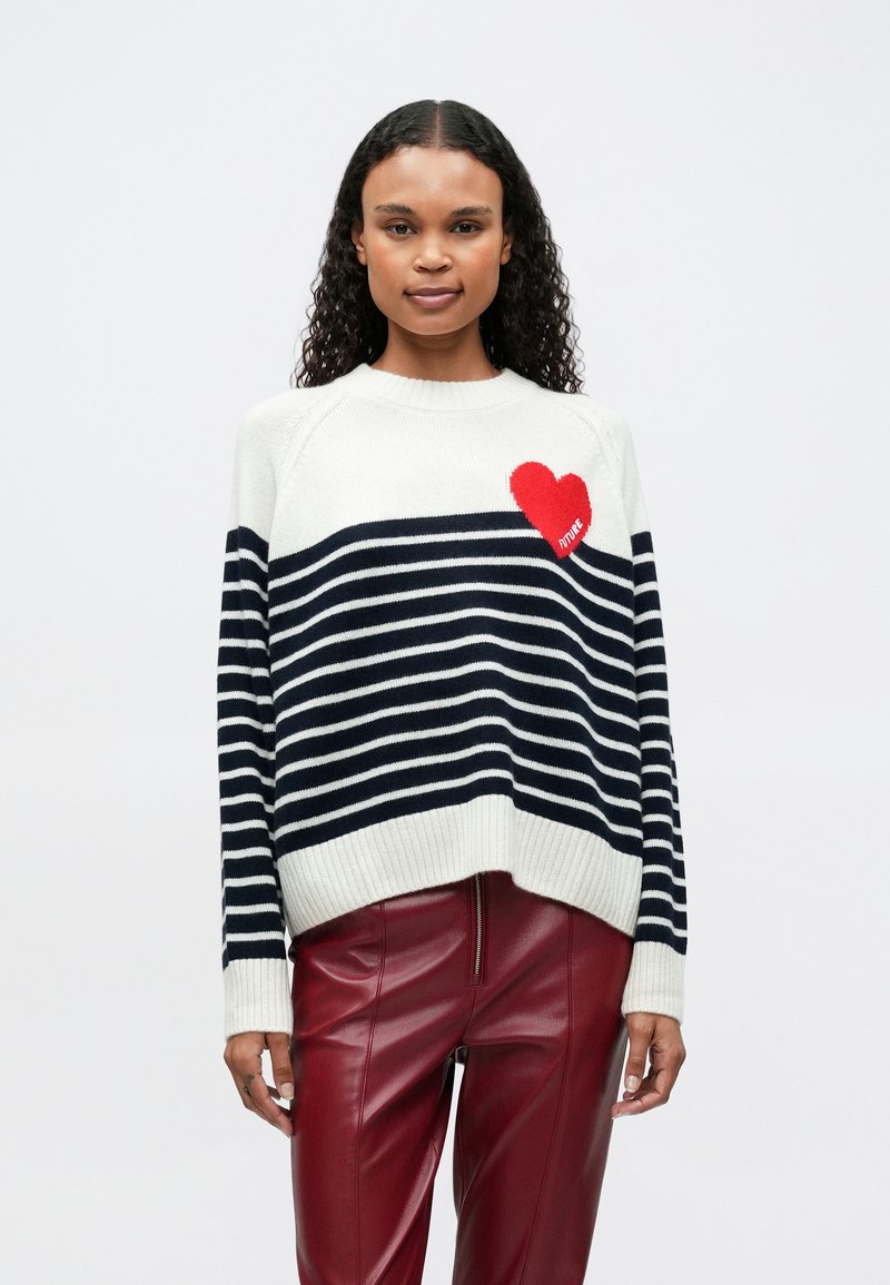 Mujer joven vistiendo un suéter a rayas blanco y azul marino con un detalle de corazón rojo y pantalones de cuero rojo, de pie contra un fondo liso.