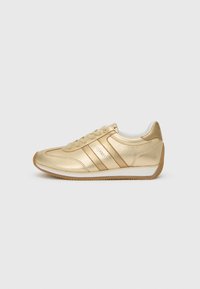 Sneaker en cuir doré avec un bout arrondi, un laçage à l'avant et des rayures sur les côtés. Il présente une semelle blanche et des accents texturés au niveau du talon.