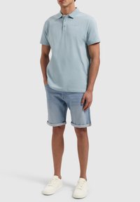 Lichtblauwe polo met drie knopen, gecombineerd met opgerolde denim shorts en witte sneakers. Zachte katoentextuur, casual pasvorm.