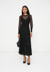 Vestido negro con mangas largas transparentes, cuerpo acanalado y falda plisada. Combinado con tacones negros y un pequeño bolso negro.