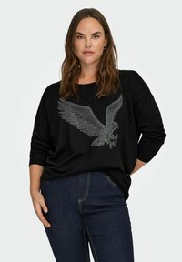 Black sweater with a loose fit, featuring a rhinestone-embellished eagle graphic on the front; paired with dark blue jeans.