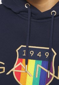 Sweat à capuche bleu marine avec un design de blason brodé doré et multicolore ; il présente des accessoires dorés sur les cordons de serrage et le texte "1949".