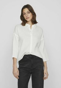 Vit skjorta med knappar i en texturerad tyg, krage och ärmar i 3/4-längd, tillsammans med svarta, högmidjade jeans.