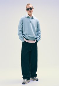 Világoskék kötött pulóver galléros nyakkal, rózsaszín ing fölé rétegezve, laza fekete farmernadrággal és szürke sportcipővel párosítva. Fekete napszemüveg.